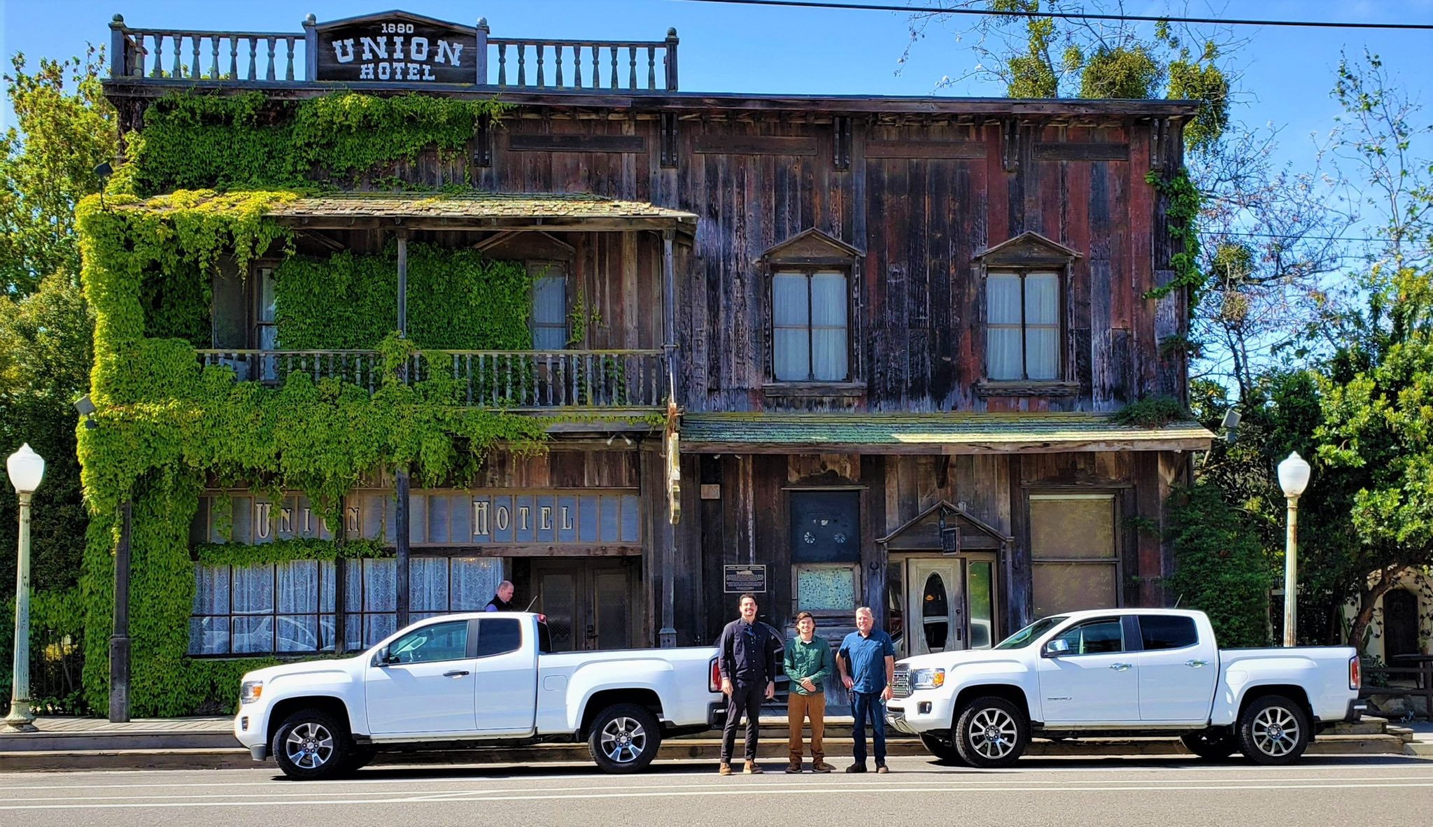 Three men pose between two white GMC pickup trucks on a sunny day. Behind them is the historic and weathered wooden Union Hotel (1880) with a distinctive ivy-covered facade and ornate lamps.